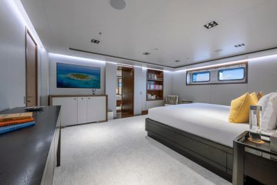 A luxurious bedroom with a large bed and a TV displaying a tropical island in the foreground, and bookshelves and windows looking out to the sea in the background.