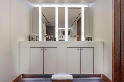 A modern bathroom vanity with a white countertop and two cabinets in the foreground is reflected in a large mirror with vertical lights in the background.