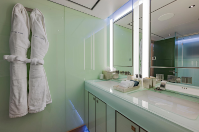 Two white bathrobes hang on a pale green wall in the foreground, while a modern bathroom vanity with a large mirror and sink is visible in the background.