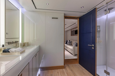 A modern bathroom with dual sinks and a glass shower stall is visible in the foreground, while a glimpse of a plush living area with a sofa and pillows can be seen through an open doorway in the background.