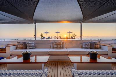 A luxurious yacht deck with comfortable seating in the foreground overlooks a serene ocean sunset with beach chairs and umbrellas in the background.