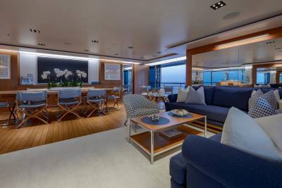 A spacious yacht interior features a dining area with a long table and chairs in the foreground, while a plush seating area with a coffee table and a view of the ocean is in the background.