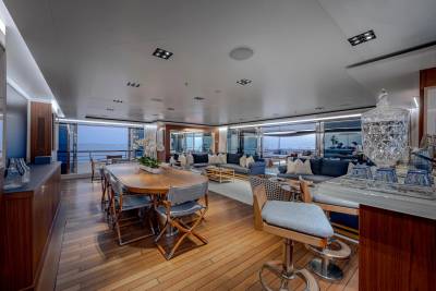 A spacious yacht interior features a wooden dining table with chairs in the foreground and a large, comfortable seating area with ocean views in the background.