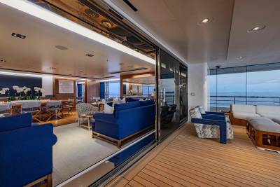 A luxurious yacht interior with a spacious deck in the foreground and a dining area and lounge visible through open sliding doors in the background.