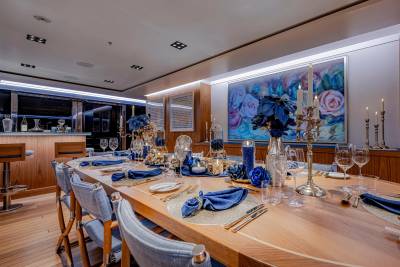 A beautifully set oval dining table with blue accents and floral arrangements is in the foreground, while a bar area and a large floral painting adorn the background.
