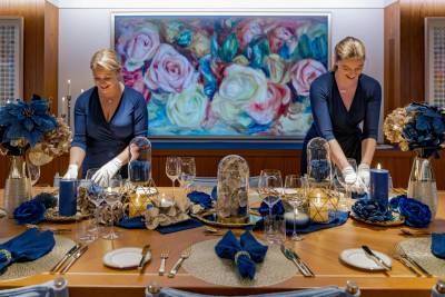Two women in blue dresses set a formal dining table adorned with blue flowers and candles, with a vibrant floral painting hanging on the wall behind them.