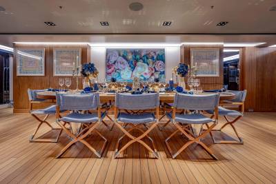 A long dining table set with blue linens and floral centerpieces is surrounded by folding chairs on a wooden deck, with a large floral painting and framed artwork adorning the wood-paneled walls in the background.