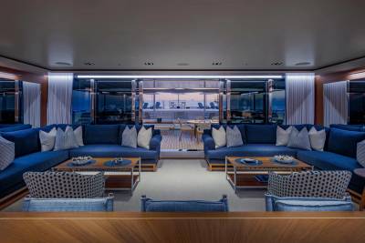 Luxurious blue sofas and patterned armchairs fill the foreground of a spacious yacht lounge, while the background reveals a serene ocean view through large windows.