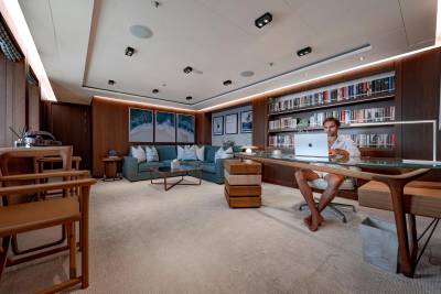 A man works on a laptop at a desk in the foreground, while a plush sectional sofa and a wall of bookshelves fill the background of a luxurious room.