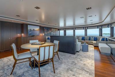 A circular dining table with chairs in the foreground is set against a spacious lounge area with a view of the ocean through large windows in the background.