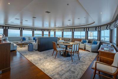 A luxurious yacht lounge with comfortable seating and a dining area in the foreground, and a panoramic view of the ocean through large windows in the background.