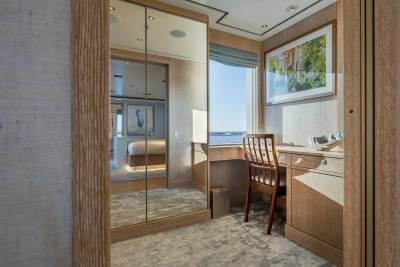 A mirrored closet door reflects a bedroom with a bird painting, while a desk with a wooden chair sits by a window overlooking the water.