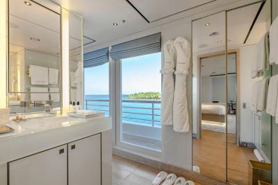 A modern bathroom with a vanity and plush robes in the foreground opens to a view of the ocean and a distant island through a large window, with a glimpse of a bedroom reflected in a mirrored wall.
