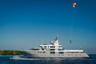 A large yacht sails on blue water with a kiteboarder in the air behind it, while palm trees line the distant shore under a clear sky.