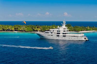 A large white yacht floats in turquoise water near a tropical island with palm trees, while a kite surfer and a jet ski are visible in the foreground.