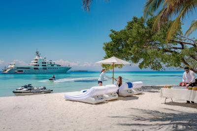 In the foreground, inflatable loungers and a table with refreshments sit on a sandy beach, while a large yacht cruises in the turquoise ocean under a clear blue sky in the background.