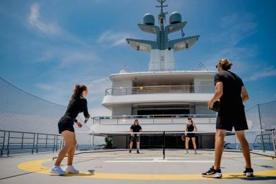 Four people play pickleball on a yacht's deck with a large white superstructure and radar in the background.