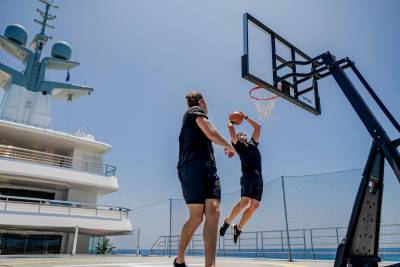 Two men play basketball on a yacht deck with a clear blue sky and ocean in the background.