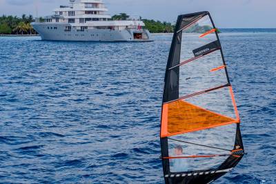 A person windsurfing in the foreground sails past a large yacht anchored near a tropical island in the background.