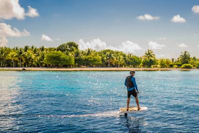 A person on a hydrofoil board glides across a bright blue ocean, with a lush green island fringed by a sandy beach and palm trees in the background under a partly cloudy sky.