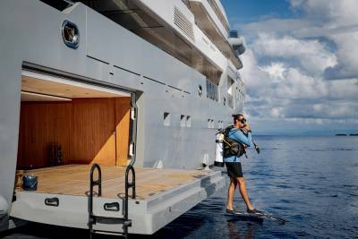 A diver in scuba gear stands on the edge of a yacht's open stern, ready to enter the calm blue ocean under a cloudy sky.