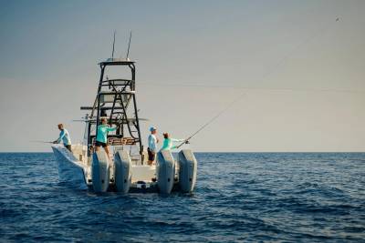 Four people fish from a large boat with triple outboard engines on a calm blue sea under a clear sky.