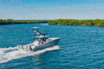 A sleek gray boat with multiple outboard motors speeds through a channel, leaving a white wake, with lush green trees lining the banks under a clear blue sky.