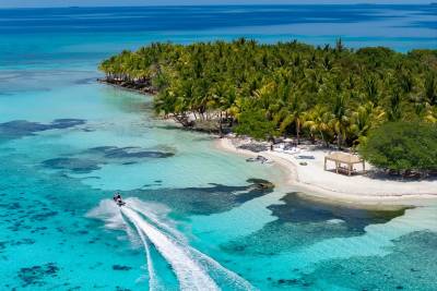 A jet ski speeds through turquoise waters in the foreground, leaving a white wake, while a lush, palm-covered island with a sandy beach and a thatched hut sits in the background under a clear blue sky.