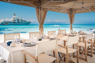 A formal dining table is set on a white sand beach with a large yacht and turquoise water in the background.