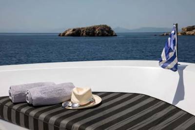 Two rolled towels, a hat, and sunglasses rest on a striped lounge chair in the foreground, with a Greek flag waving beside a rocky island and the calm blue sea in the background.