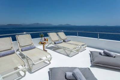 Four lounge chairs and two large floor cushions are arranged on a white deck, with a small table holding drinks and snacks, all set against a backdrop of the deep blue sea and a clear sky.