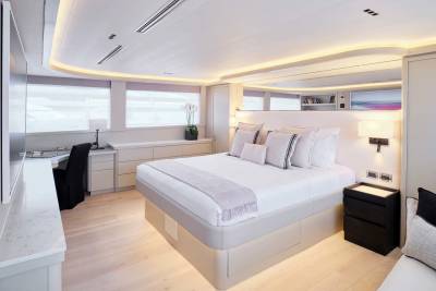 A luxurious yacht bedroom features a plush bed in the foreground and a desk area with large windows in the background.
