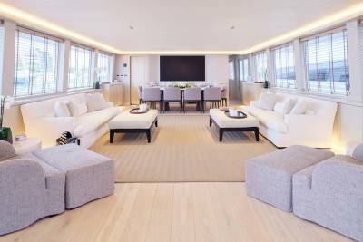 Plush white sofas and ottomans fill the foreground of a spacious, well-lit yacht interior, while a long dining table with chairs and a large screen are visible in the background.