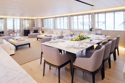 A formal dining table set for a meal is in the foreground, while a spacious, well-lit lounge area with sofas and ottomans fills the background.