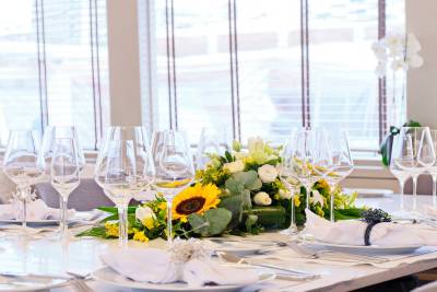 A beautifully set dining table with a vibrant floral centerpiece and wine glasses in the foreground is illuminated by natural light streaming through a window in the background.