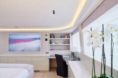 A modern bedroom features a large abstract artwork on the wall above a dresser in the foreground, while a built-in desk with a bookshelf and a window with white orchids fill the background.