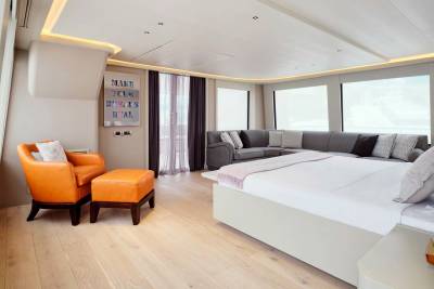 An orange armchair and ottoman sit in the foreground of a spacious room with a large bed and sectional sofa, all illuminated by natural light from large windows in the background.