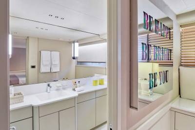 A modern bathroom with a white vanity and sink in the foreground is reflected in a mirror displaying colorful text against a background of blinds and a cushioned bench.