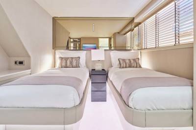 Two neatly made twin beds with decorative pillows flank a nightstand with a lamp, while a mirrored wall reflects the room and blinds cover a window in the background.