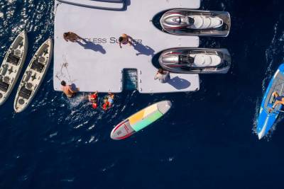 People are enjoying various water activities like kayaking, paddleboarding, and swimming around a floating platform with two sleek boats docked, all set against a deep blue ocean.