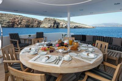 A lavish breakfast spread is set on a round wooden table on a yacht's deck, with a rocky coastline and blue sea in the background.
