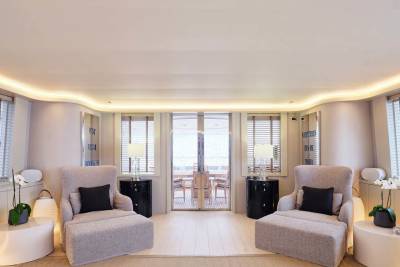 Two plush armchairs with black accent pillows sit in the foreground of a bright room with large windows and a balcony in the background.