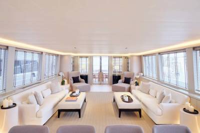 Plush white sofas and armchairs fill the foreground of a bright, modern yacht salon, while large windows with blinds reveal a glimpse of the outdoors in the background.