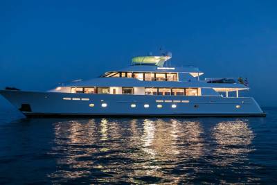 A luxurious white yacht with illuminated windows floats on calm, dark water, reflecting its lights against a deep blue twilight sky.