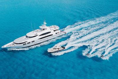 A large white yacht cruises through turquoise waters, trailed by a smaller boat and two jet skis.