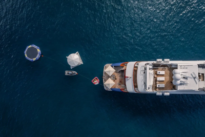 A large yacht is anchored in deep blue water, with a towable inflatable, a small dinghy, and a floating umbrella in the foreground.