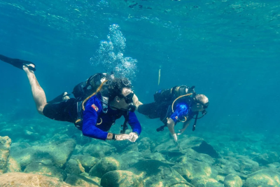 Two divers swim over a rocky seabed in clear blue water, with sunlight filtering from above.