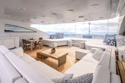 A spacious, white-cushioned outdoor lounge area with a wooden coffee table in the foreground overlooks a vast blue ocean under a cloudy sky in the background.
