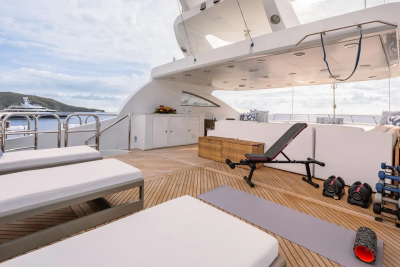 Two white lounge chairs and a yoga mat with weights are on the wooden deck of a yacht, with a cloudy sky and distant islands in the background.