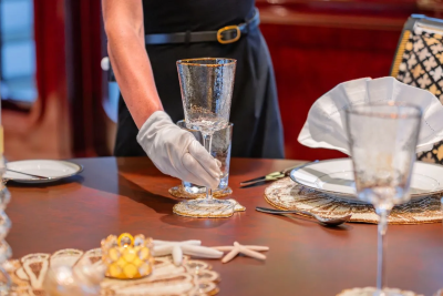 A gloved hand places stacked glasses on a table set with elegant dinnerware, while a richly decorated chair and red drapery form the background.